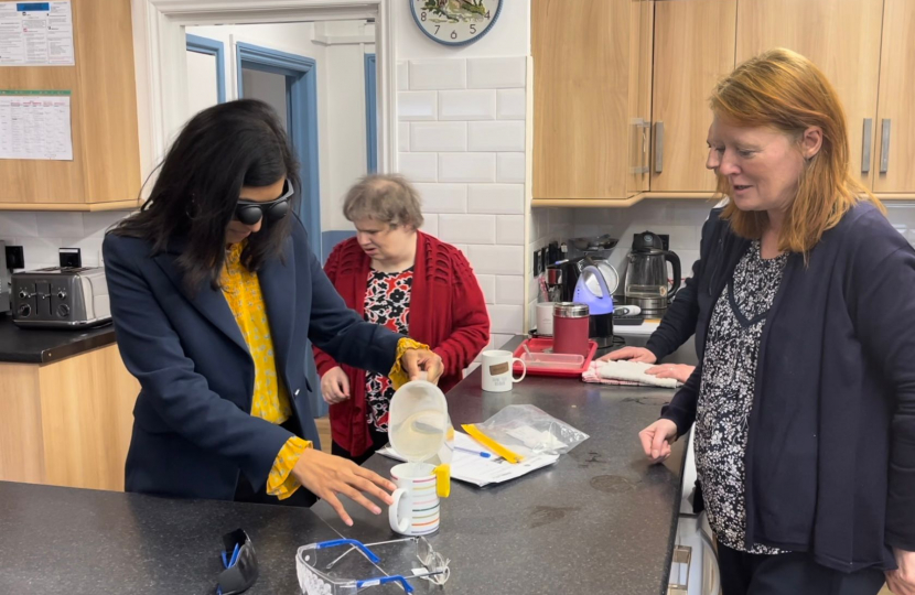 Claire is wearing glasses that simulate visual impairment, pouring water into a jug using a device that beeps when the cup is full.