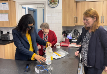 Claire is wearing glasses that simulate visual impairment, pouring water into a jug using a device that beeps when the cup is full.